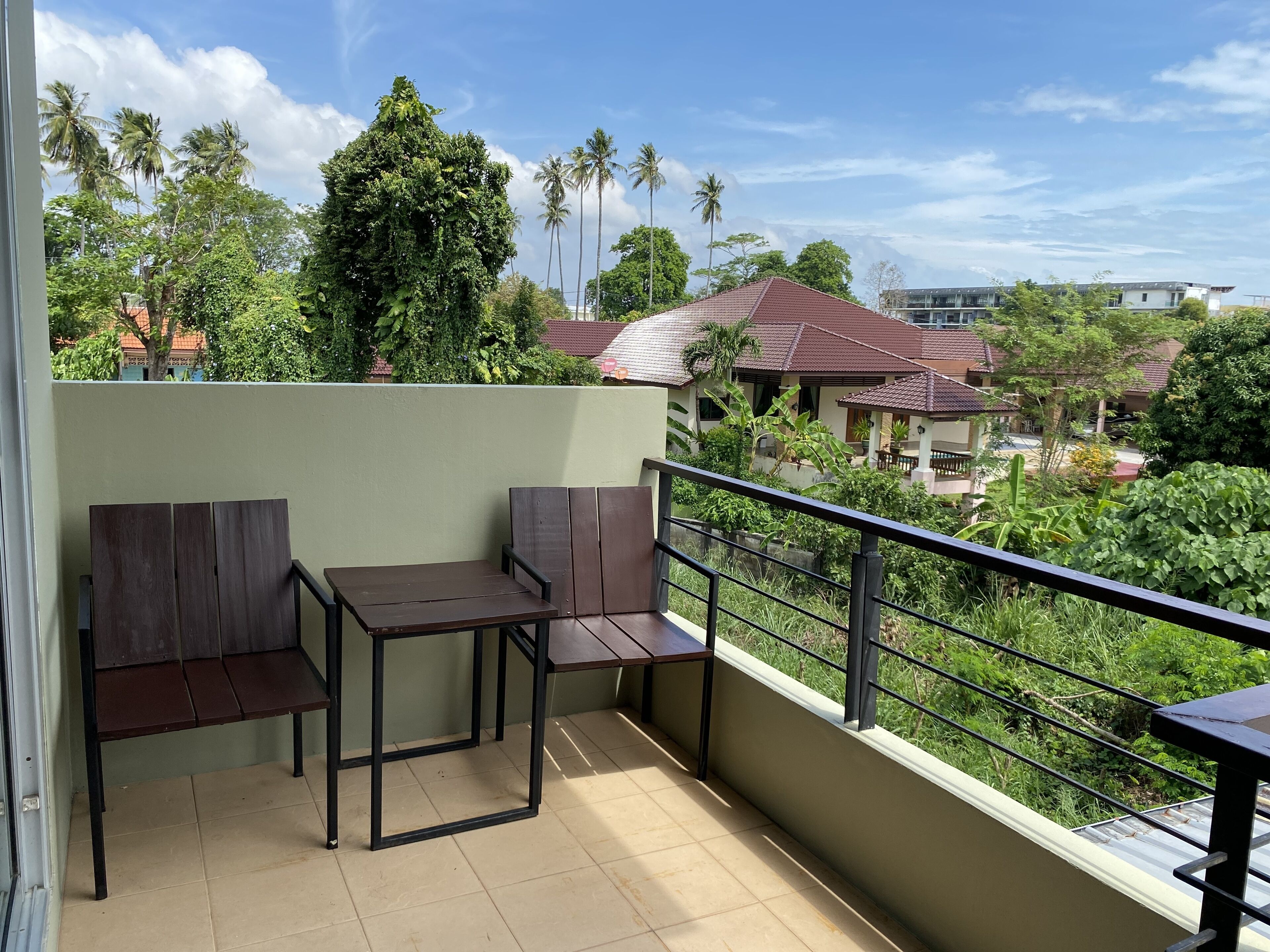 A balcony with two wooden chairs and a small table, overlooking a residential area with houses and palm trees.