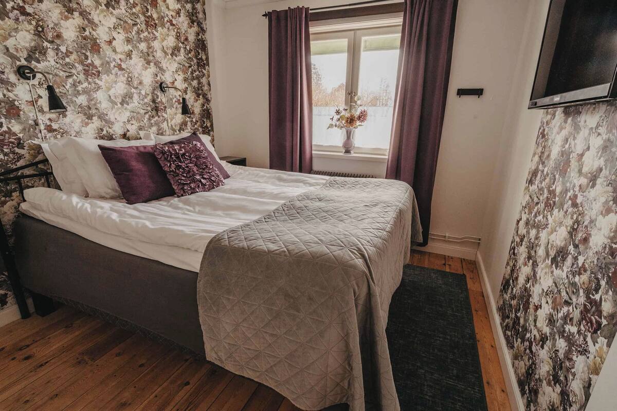 Un dormitorio con una cama, una mesita de noche, una ventana con cortinas y un papel tapiz con motivos florales.