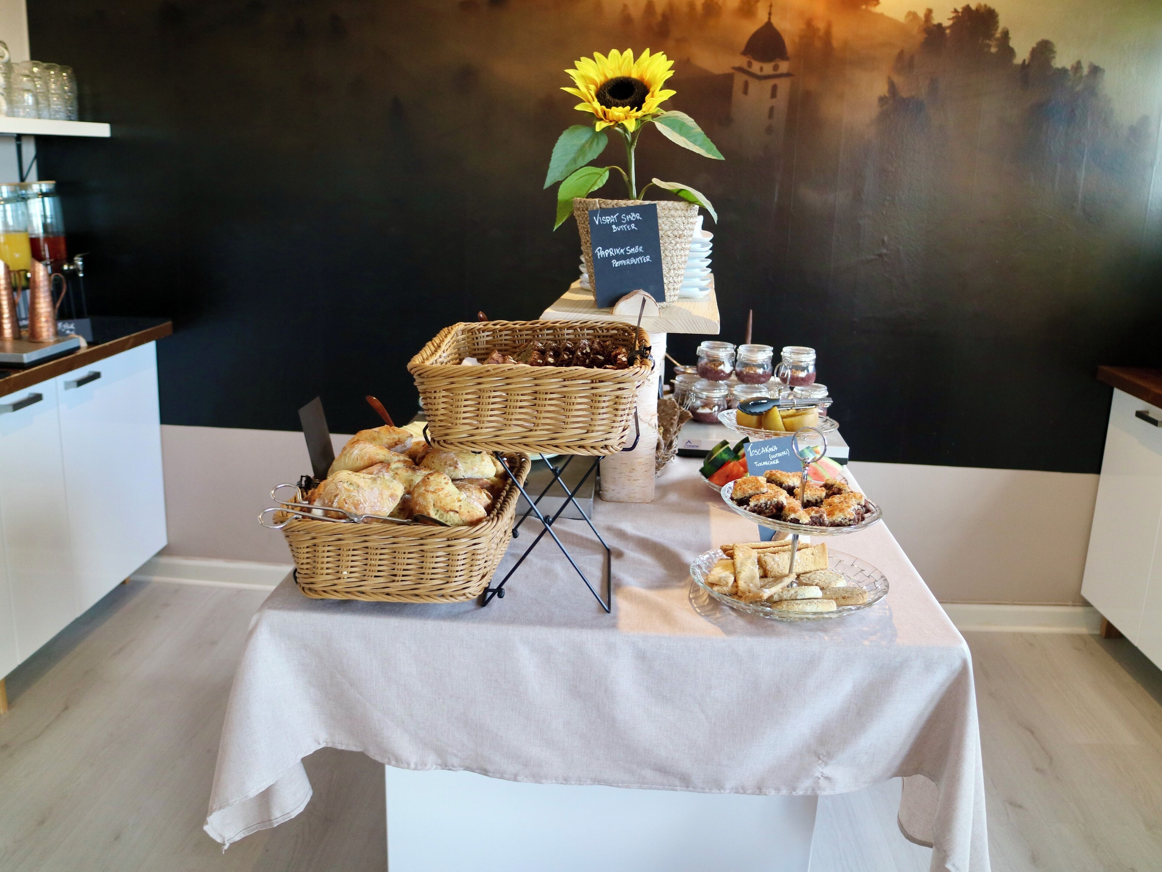 A buffet table with baskets of pastries, a sunflower centerpiece, and a scenic mural in the background.