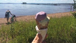 Une personne tenant une glace à la main, près d’un lac, avec un vélo et une jetée visible au loin.