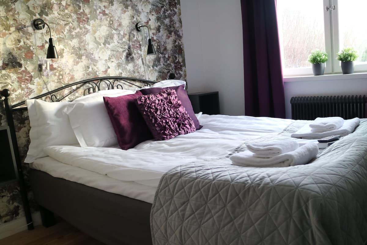Una cama bien hecha con sábanas blancas y una colcha gris acolchada, un cojín decorativo morado y dos pequeñas macetas con plantas en el alféizar.