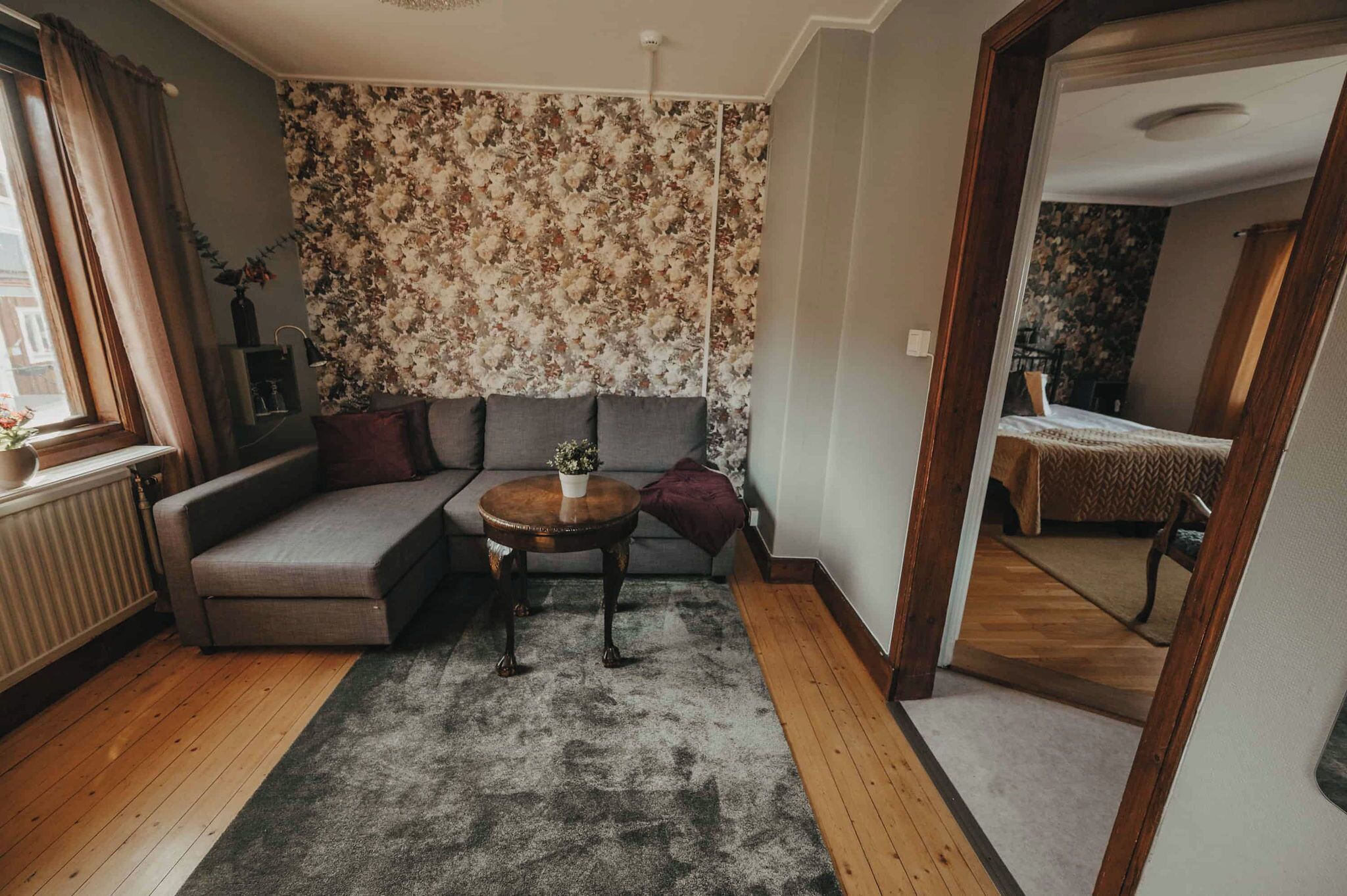 Een gezellige woonkamer met een grijze hoekbank, een ronde houten salontafel en behang met een bloemenpatroon.