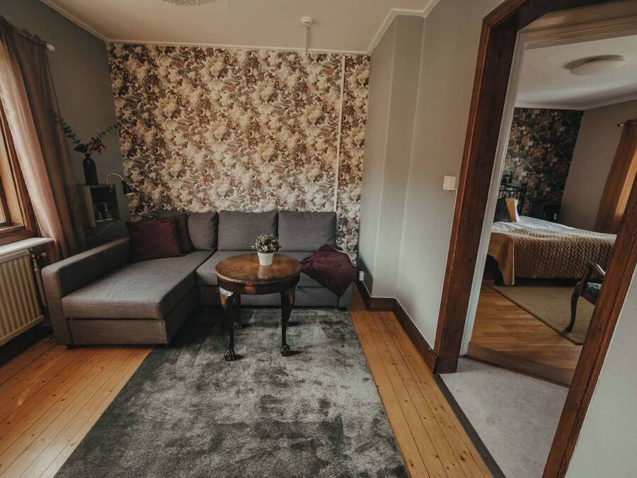 Een gezellige woonkamer met een grijze hoekbank, een ronde houten salontafel en behang met een bloemenpatroon.