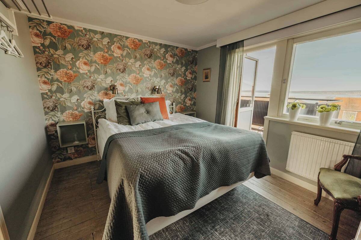 Un dormitorio con papel tapiz floral, una cama con manta verde, una mesita de noche, una silla y una ventana con vistas al exterior.