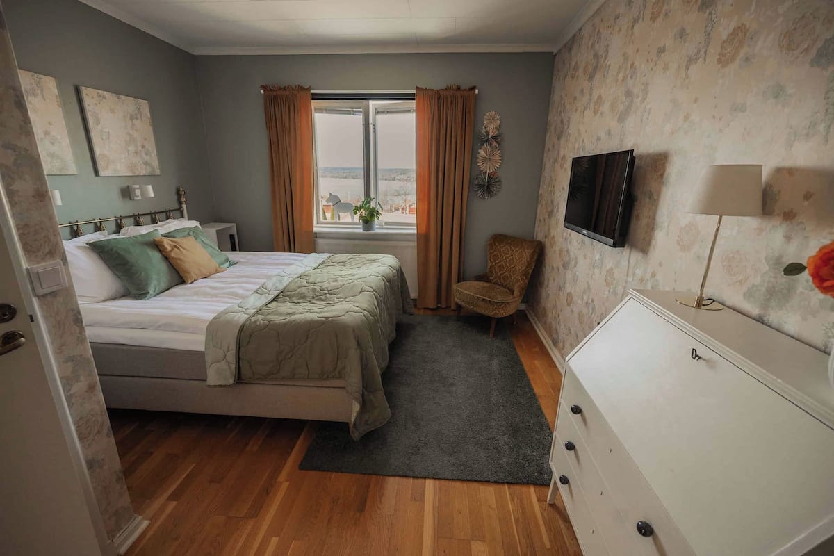 Un dormitorio con cama, cómoda, silla, ventana con cortinas y televisor en la pared.