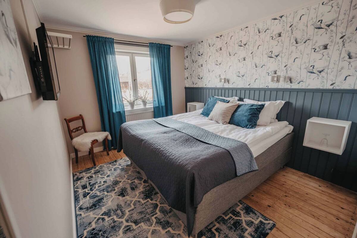 Un dormitorio con cama, una silla, un ventanal con cortinas, un televisor instalado en la pared y un papel tapiz decorado.