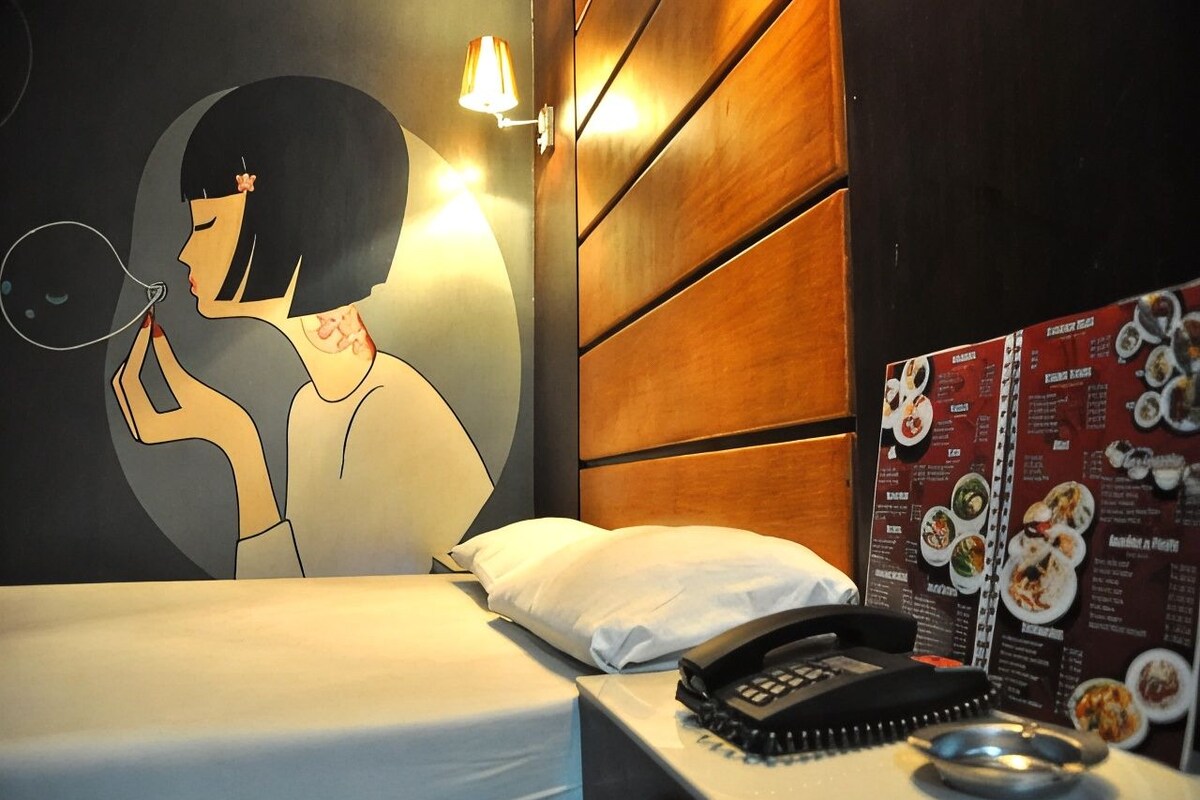 Une chambre d’hôtel équipée d’un lit, d’un téléphone fixé au mur, d’un tableau affichant le menu et d’une œuvre d’art décorative.