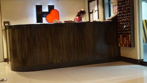 Een receptiebalie met een donkere houtafwerking, een groot, oranje hartvormig bord erboven, en een muur met verschillende mededelingen en een presentatie van rode en gele objecten.