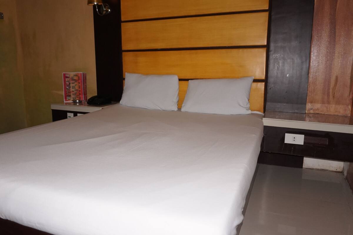 Een keurig opgemaakt bed met witte lakens, een houten hoofdbord, een nachtkastje met een lamp en een klein nachtkastje met een telefoon.