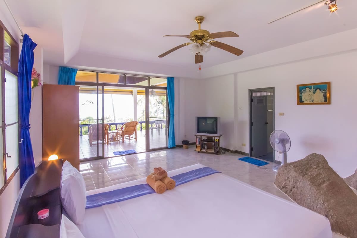 A hotel room with a bed, a ceiling fan, a television, and a balcony with chairs.