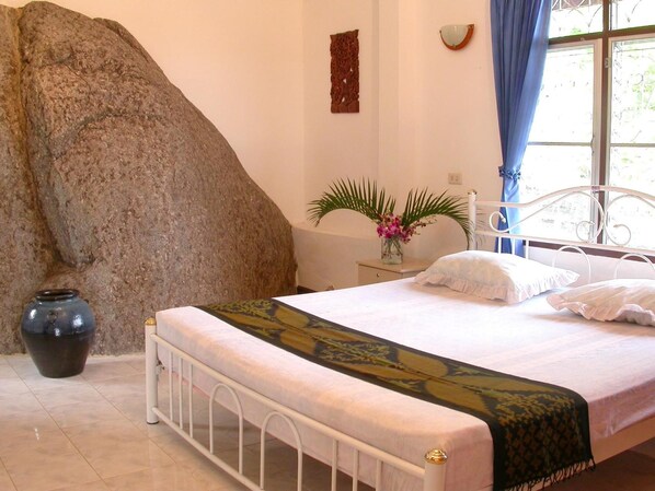 Un dormitorio con una gran formación rocosa, una cama con colcha estampada, un jarrón con flores y una ventana con cortinas.