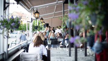An outdoor seating area with tables and chairs, a covered roof, hanging plants, and a street view in the background.