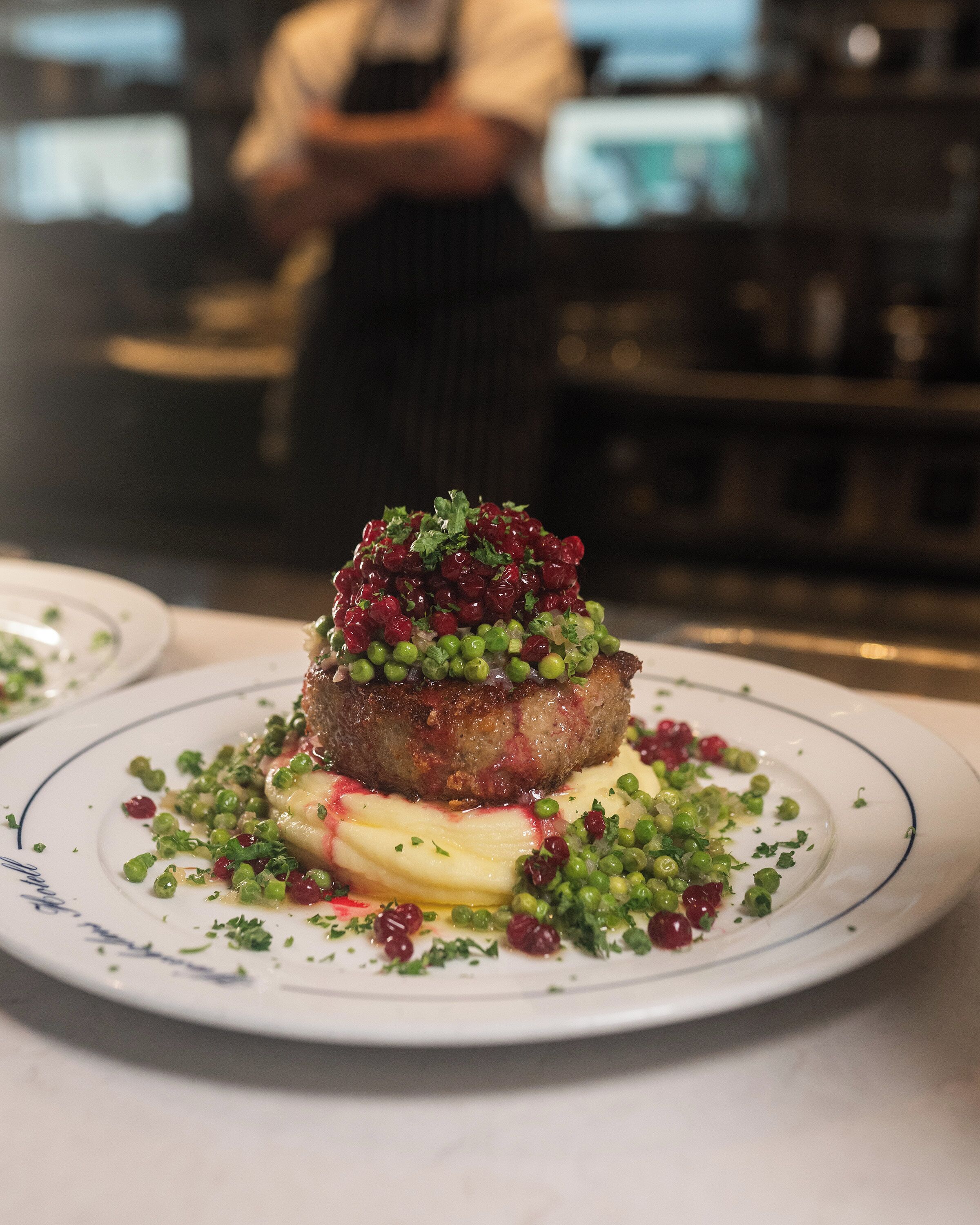 Un plato con un guiso de carne como pieza central, adornado con guisantes y hierbas, sobre un plato blanco con un borde azul.