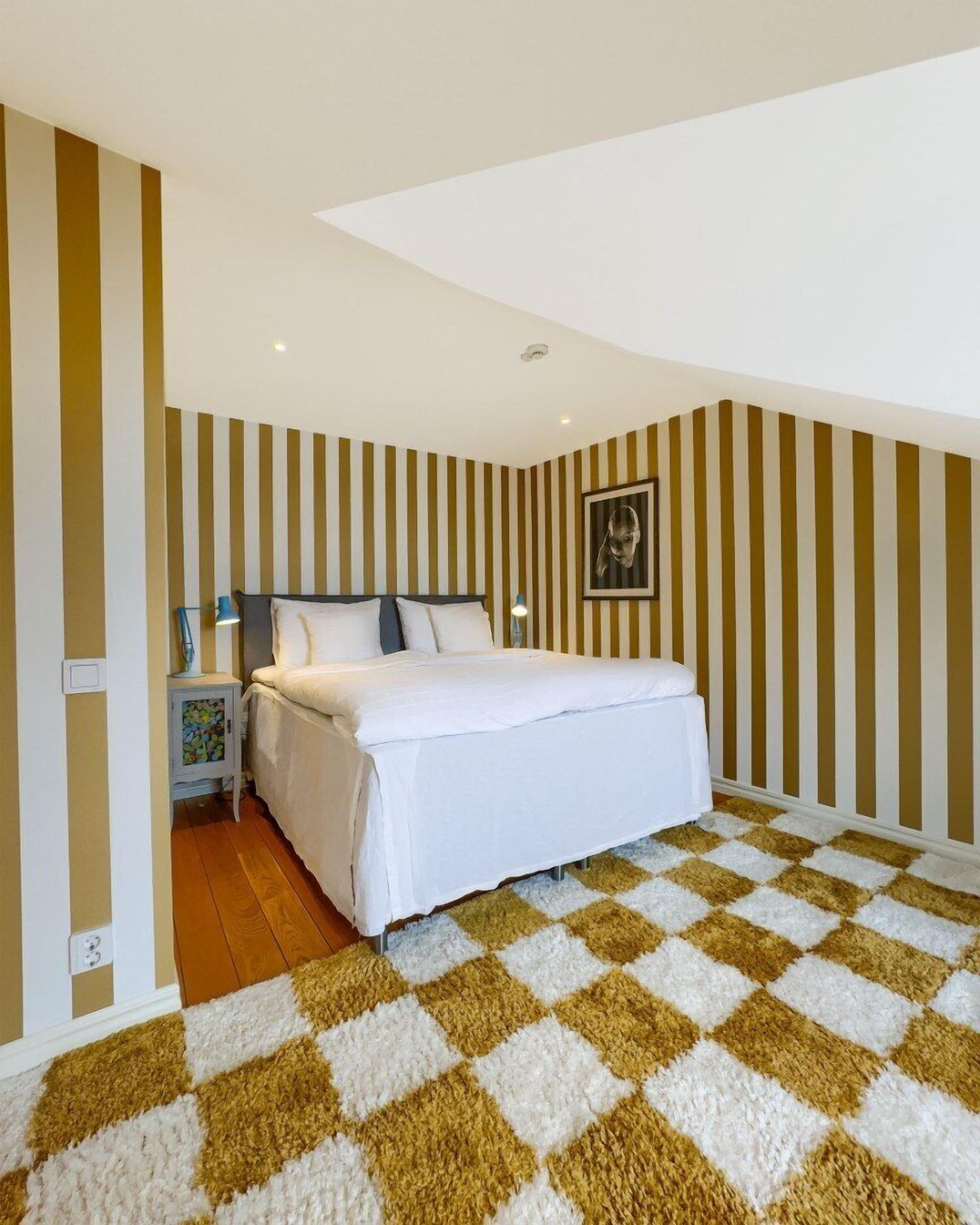 A bedroom with a bed, a nightstand, a wall-mounted lamp, and a checkered rug.