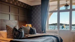 A bedroom with a large bed, patterned pillows, a view of the water through a window, and a decorative wall panel.