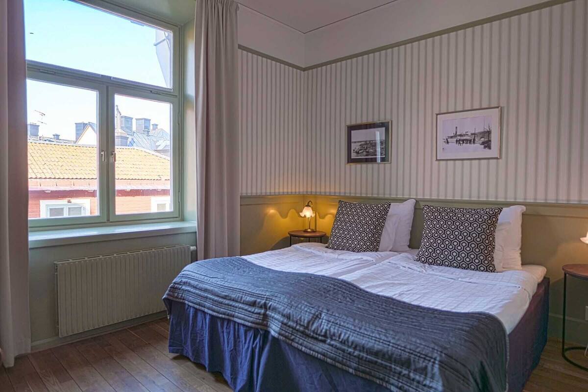 A hotel room with a large bed, a window with curtains, a radiator, and two framed pictures on the wall.