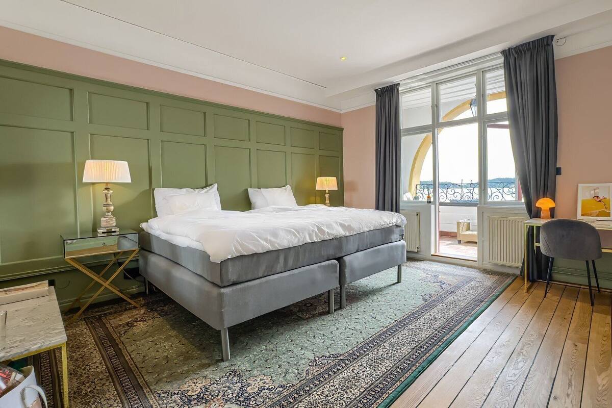 A hotel room with a bed, two bedside lamps, a desk with two chairs, and a view of the outside.