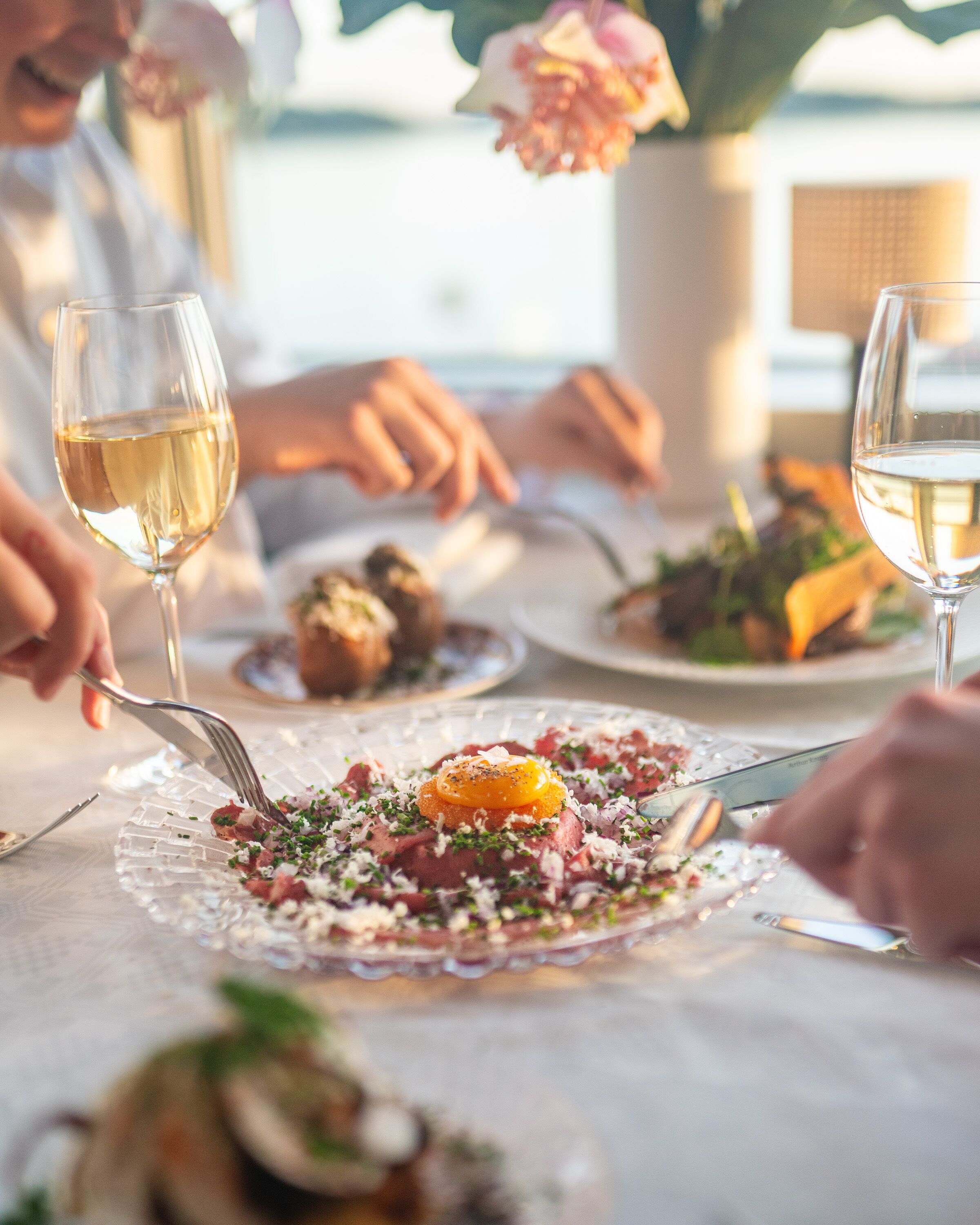 A dining table with a glass plate of food, a glass of white wine, and a fork.