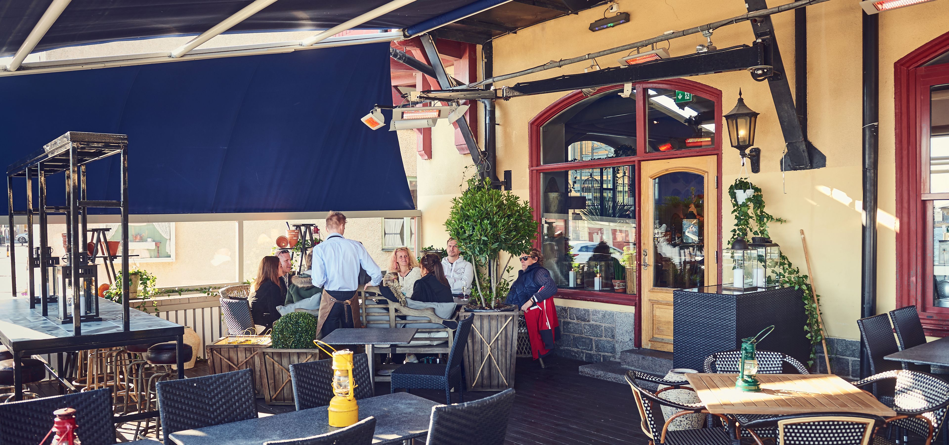 An outdoor cafe with tables and chairs, a blue awning, and a wooden deck.