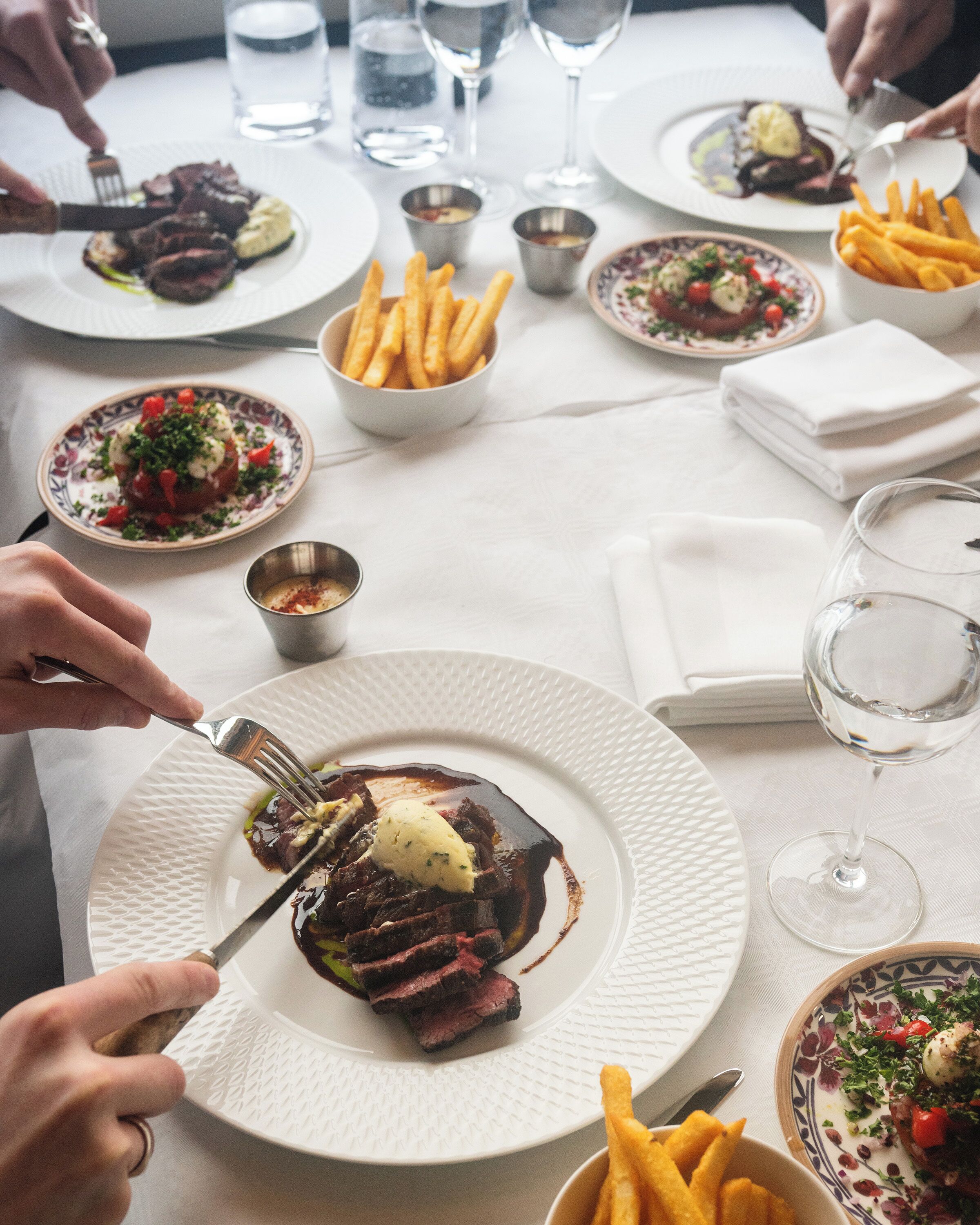 Una mesa de comedor con platos de comida, vasos de agua y una persona cortando carne.
