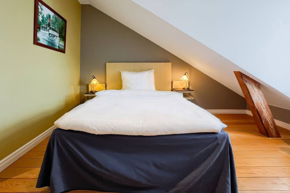 A bedroom with a bed, two bedside tables with lamps, a wooden chair, a picture on the wall, and a sloped ceiling.