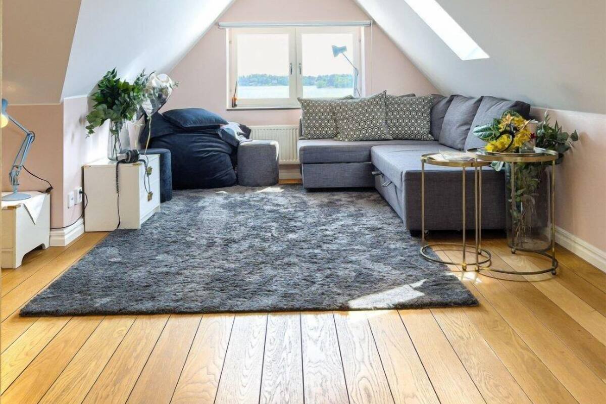 A cozy attic bedroom with a sloped ceiling, a large window, a grey sofa, a small round table, and a grey rug.