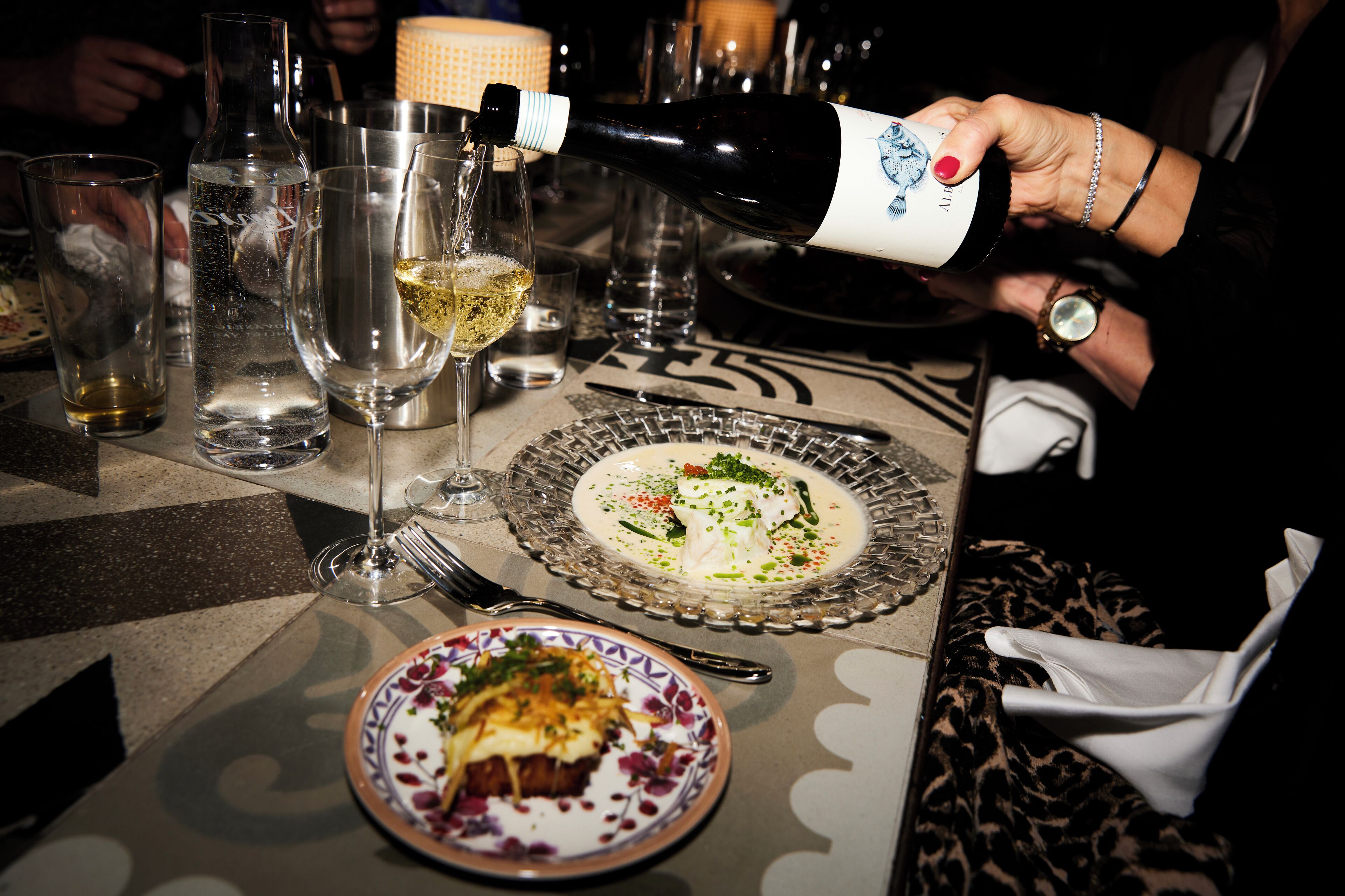 A dining table with plates of food, glasses, and a bottle of wine being poured.