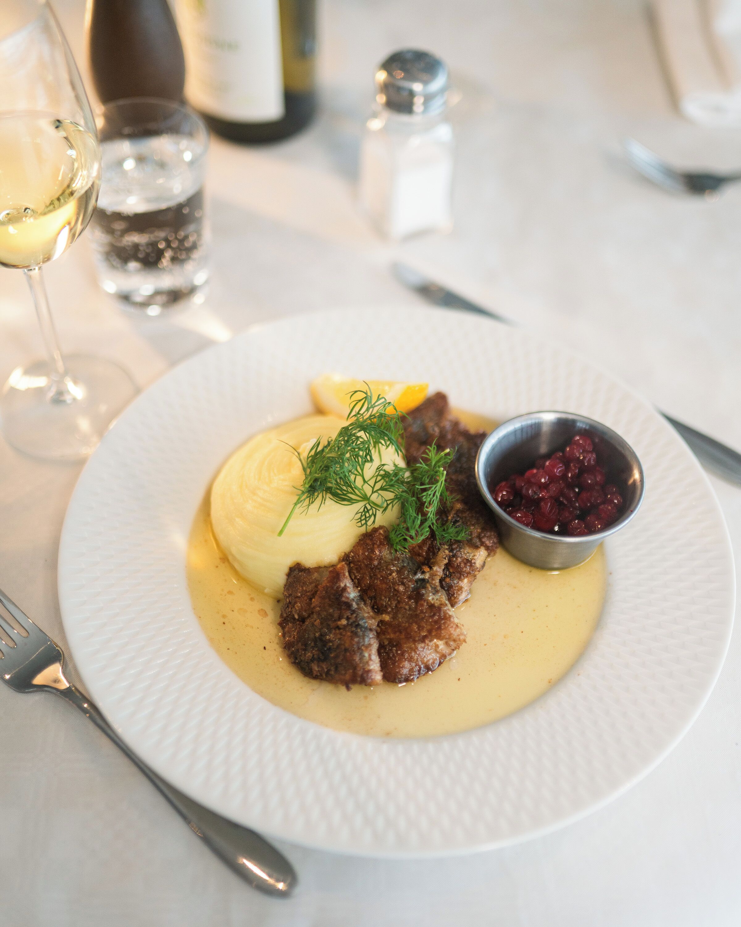 A plate of food with a creamy sauce, a small bowl of berries, and a glass of white wine on a table.