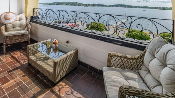 A balcony with a view of a lake and distant mountains, furnished with wicker furniture and a glass table.