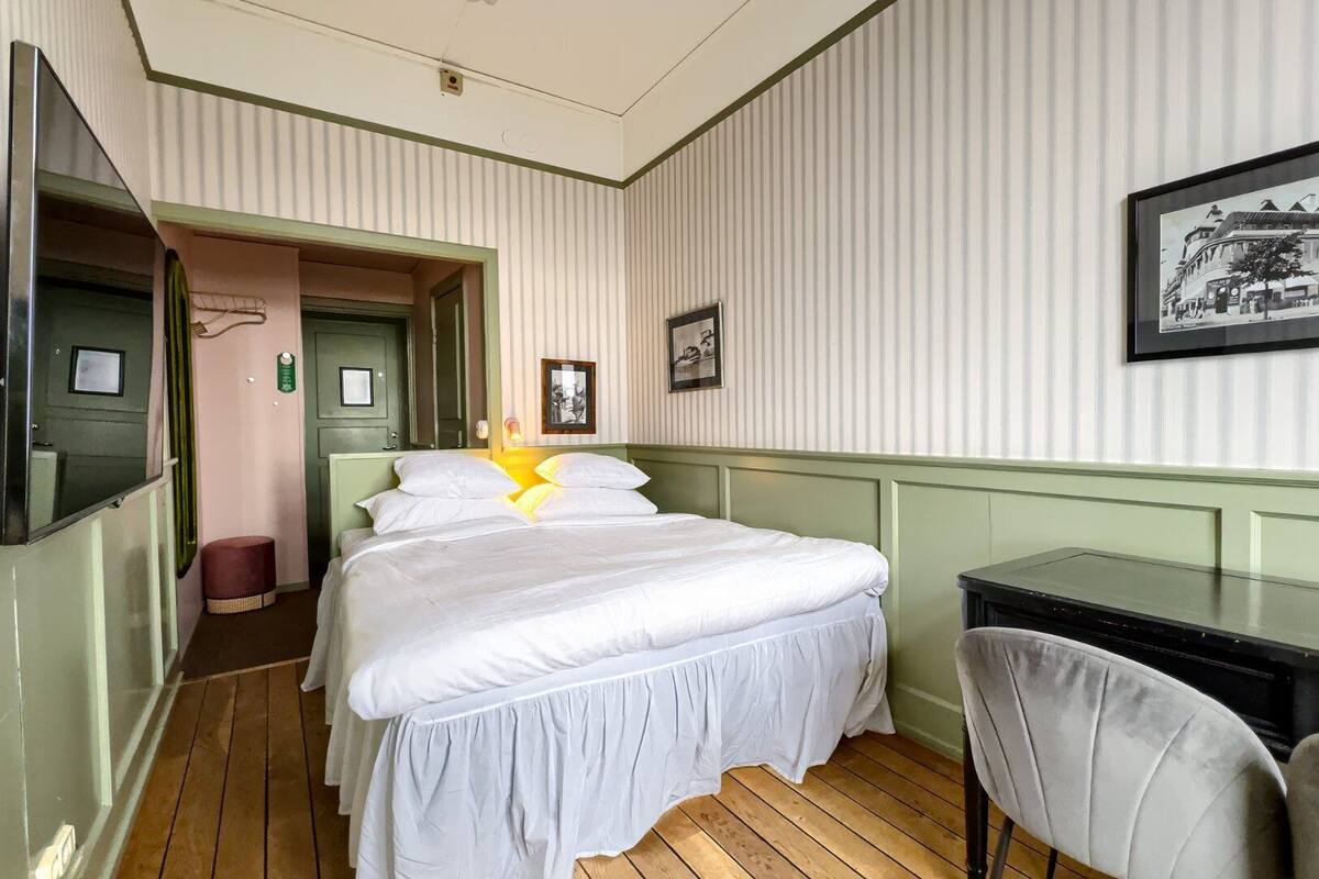A hotel room with a bed, a desk, and a chair. There is a framed picture on the wall and a door leading to another room.