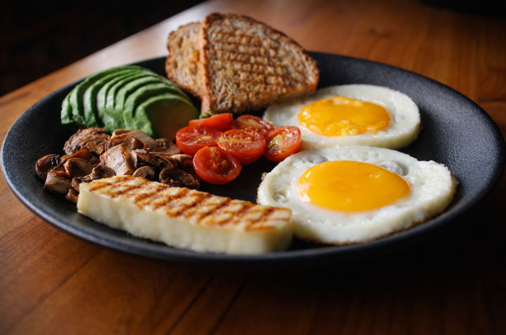 A plate with grilled meat, avocado slices, grilled tomatoes, fried eggs, and toasted bread.