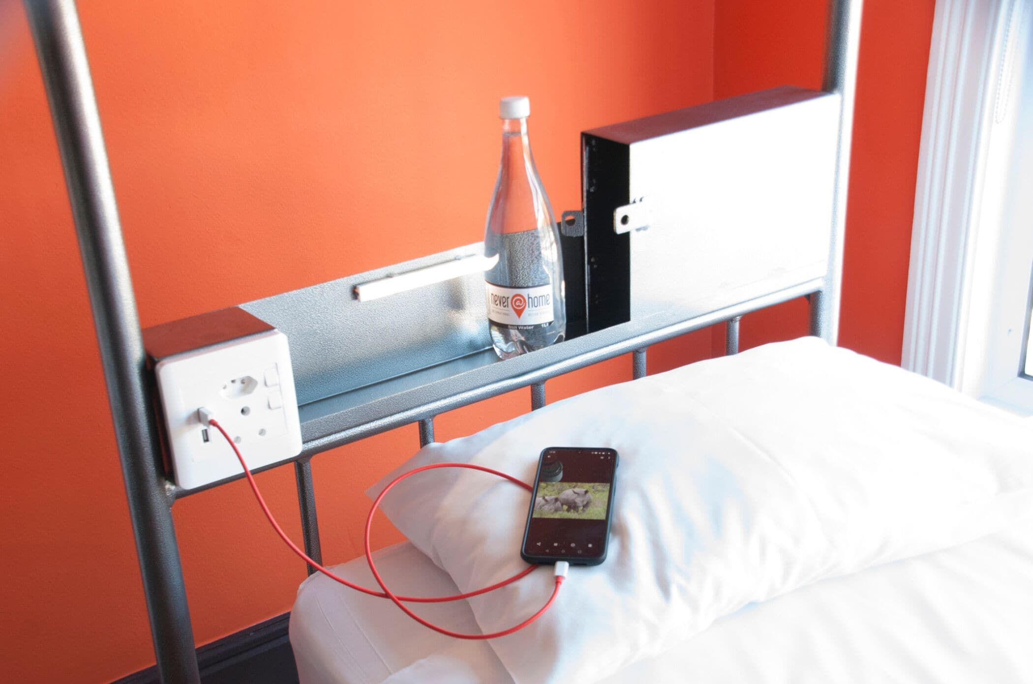 A hotel room with a bed, a bedside table with a bottle of water, a power outlet, and a smartphone charging on the bed.
