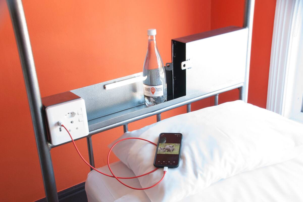 A hotel room with a bed, a bottle of water, a mobile phone, and a power outlet on the headboard.