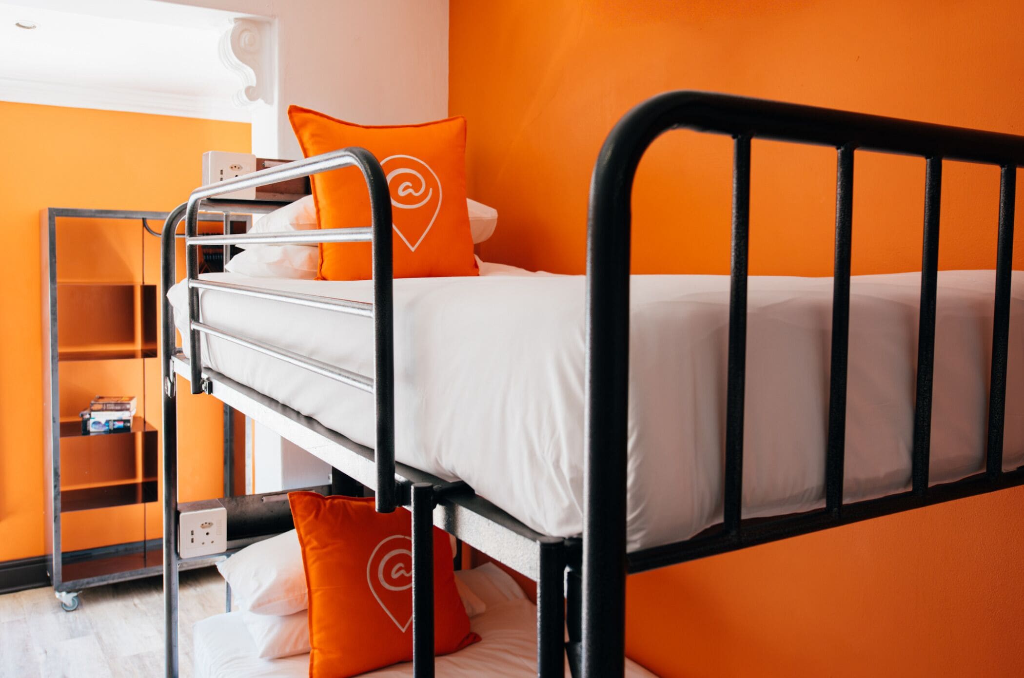 A bunk bed with orange bedding and pillows, a metal ladder, and a small shelf unit with a box on it.