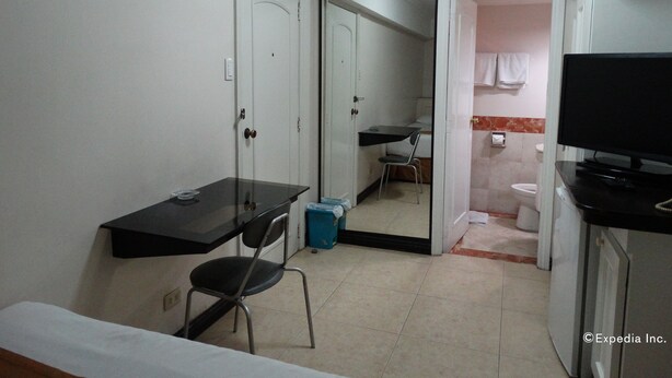 A small, compact room with a bed, a desk, a chair, a television, and a bathroom visible through an open door. - Ralph Anthony Suites (Manila)
