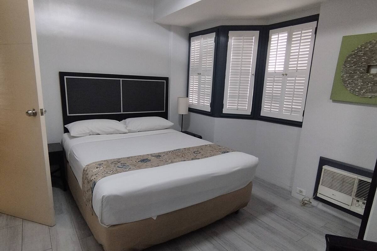 A bedroom with a bed, a nightstand, a window with shutters, a radiator, and a door.