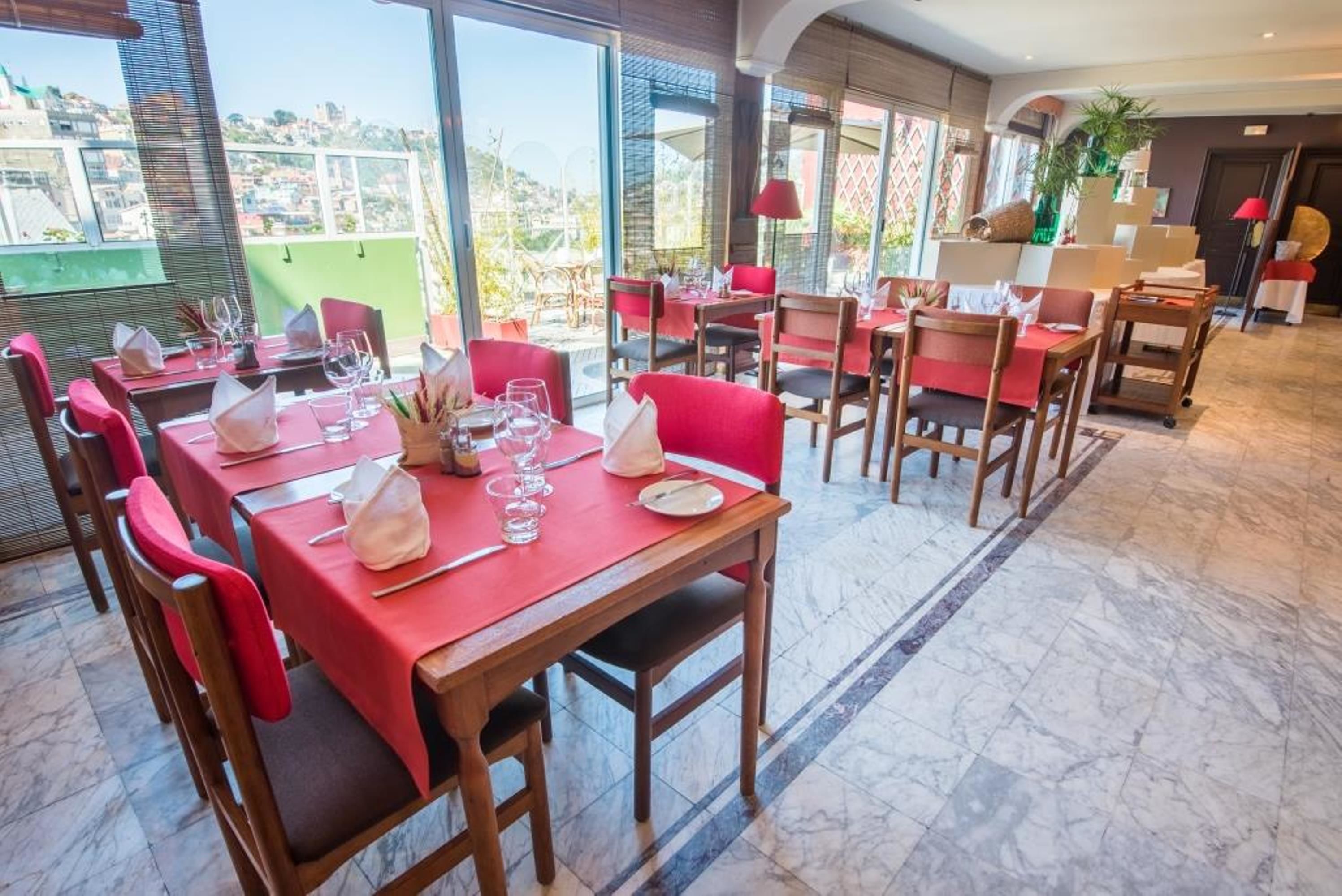 A restaurant with red tables and chairs, large windows offering a city view, and a tiled floor.