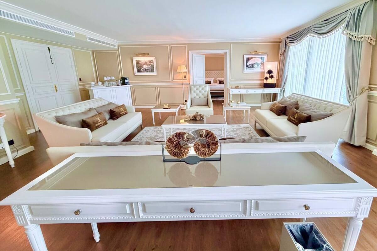 Ein geräumiges Wohnzimmer mit einem großen Glastisch, zwei weißen Sofas mit dekorativen Kissen, einer weißen Kommode und Blick auf die Küche und den Essbereich durch einen offenen Durchgang.