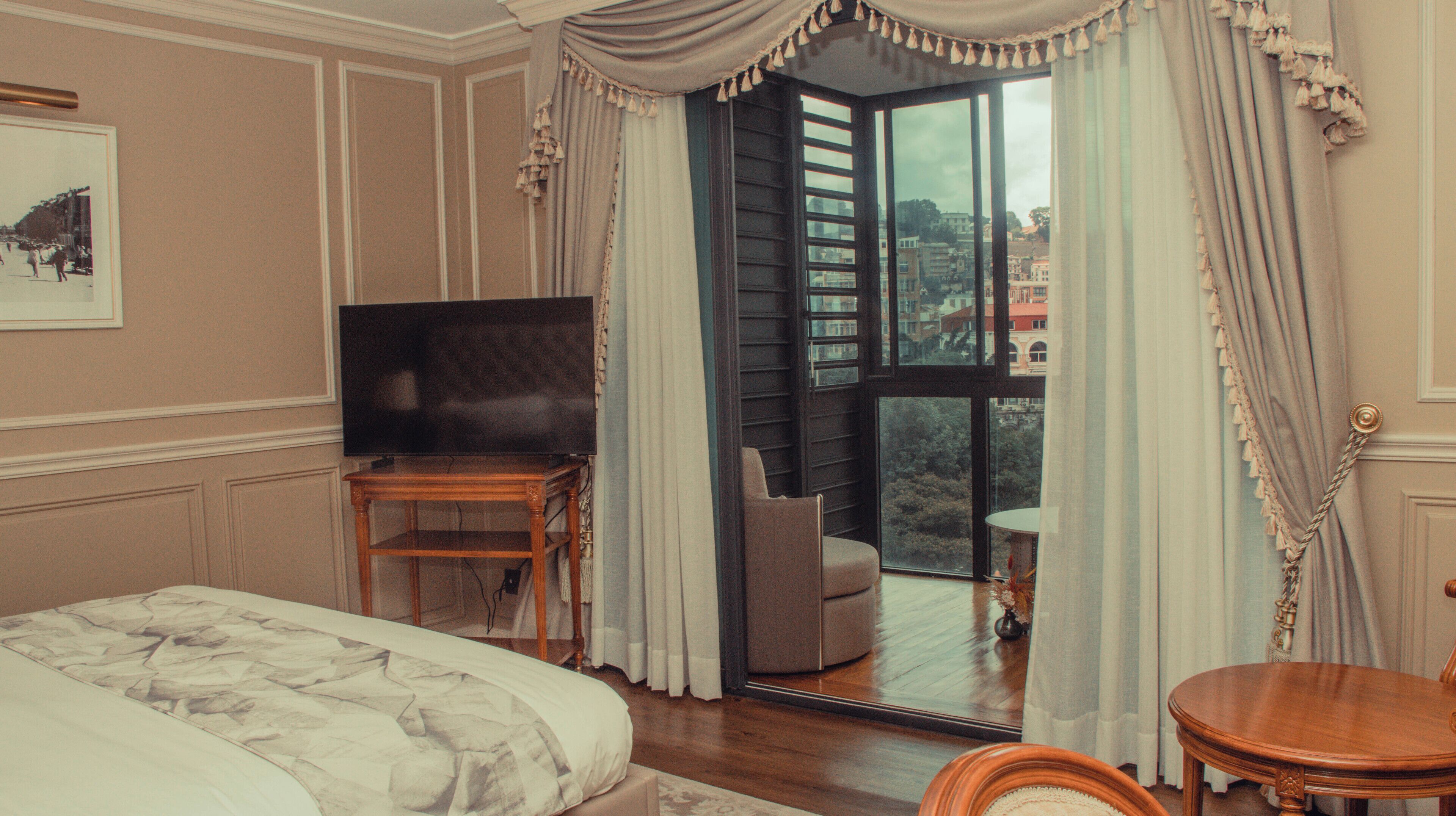 Ein Hotelzimmer mit Bett, Fernseher auf einem Holzboden, einem Balkon mit Stuhl und Blick auf Gebäude.