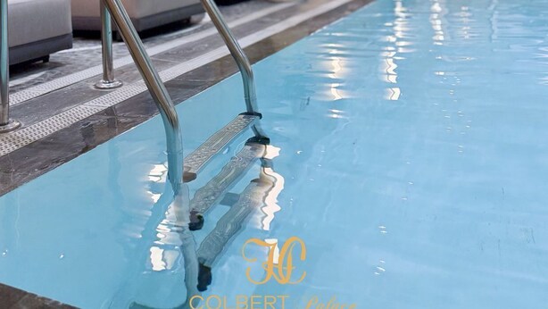 Piscina com escada de metal, cadeiras de descanso e iluminação embutida na parede.