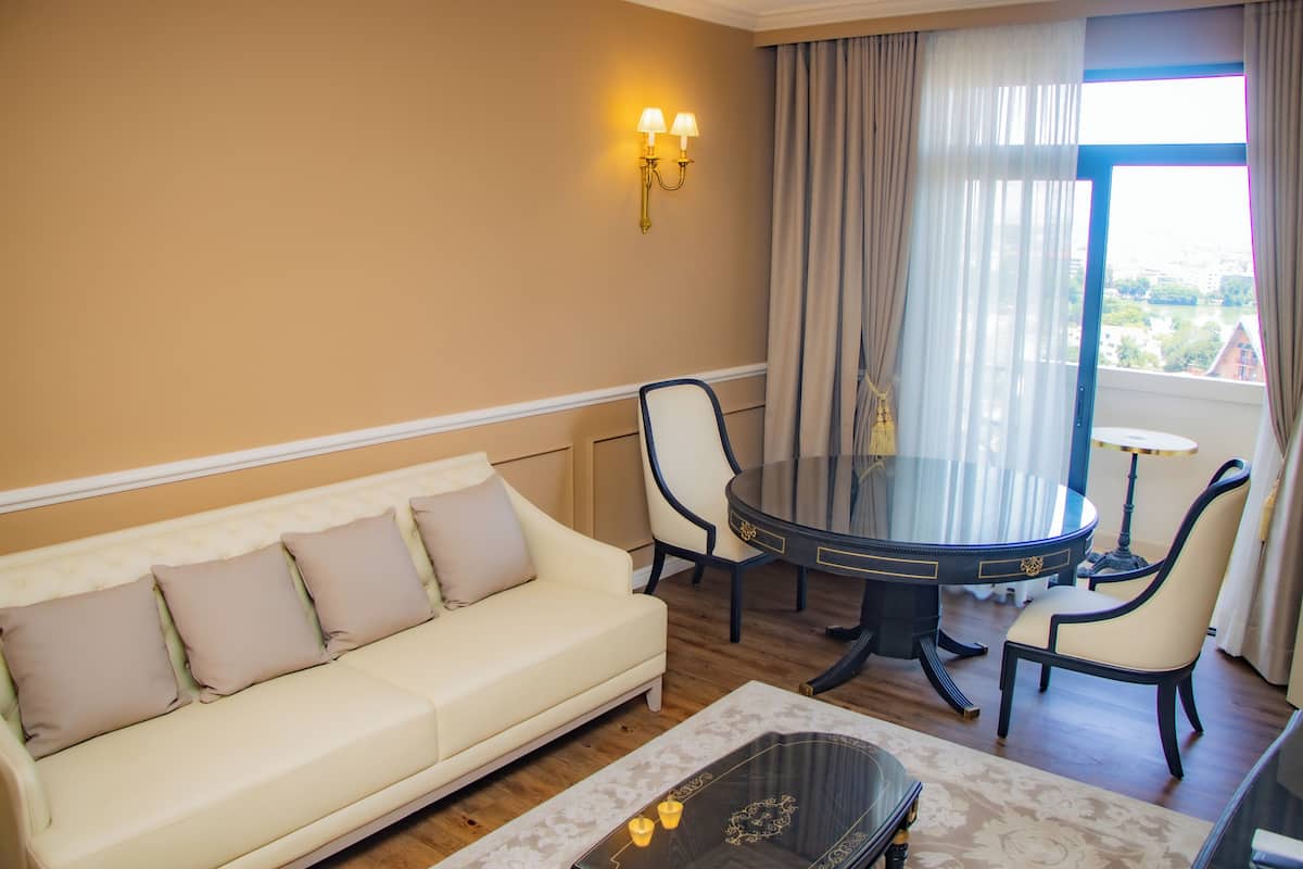 Ein Hotelzimmer mit einem beigefarbenen Sofa, einem runden Esstisch mit Stühlen, einem Couchtisch und Blick durch ein großes Fenster.