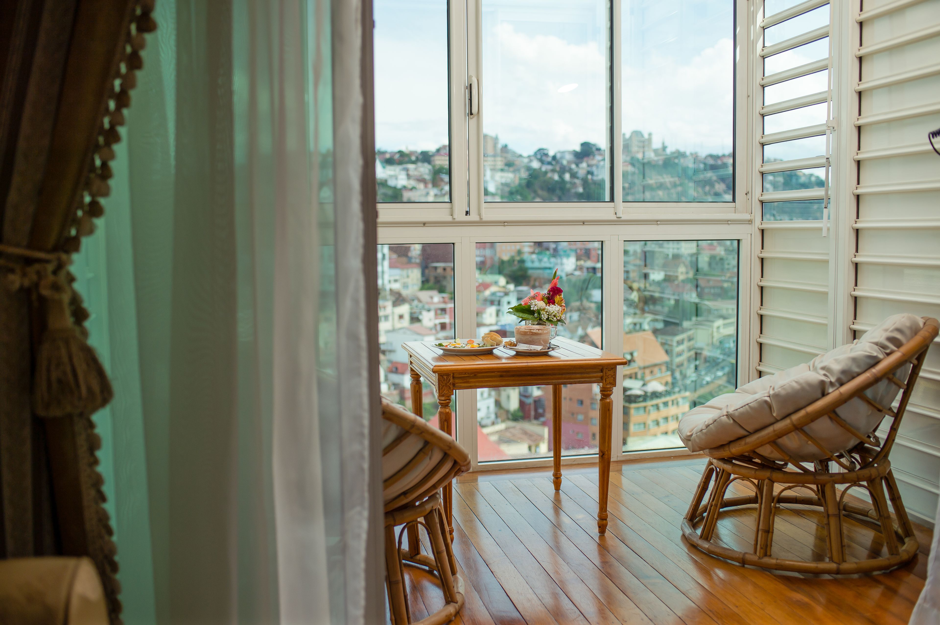 A balcony with wicker chairs, a table with a floral arrangement, and a city view through large windows.