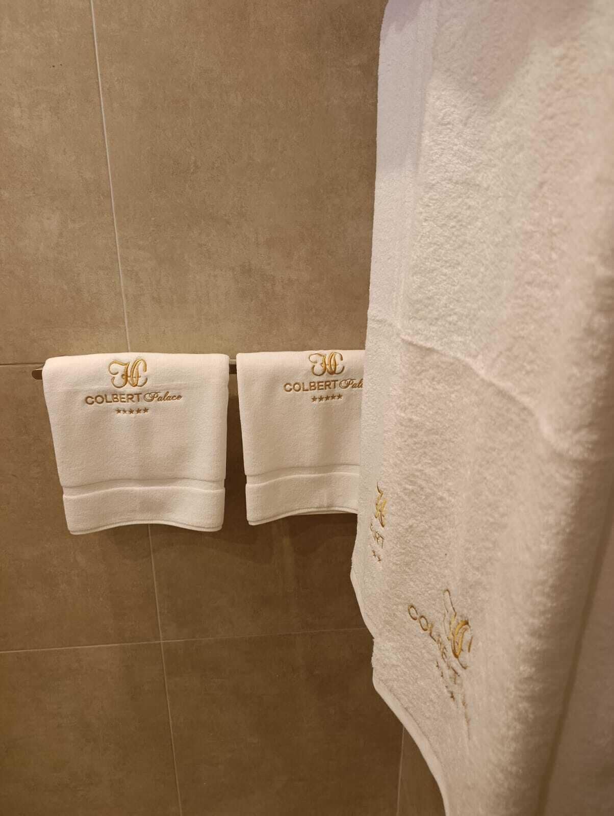 Two white towels hanging on a tiled wall with 'COLBERT Palace' embroidered on them.