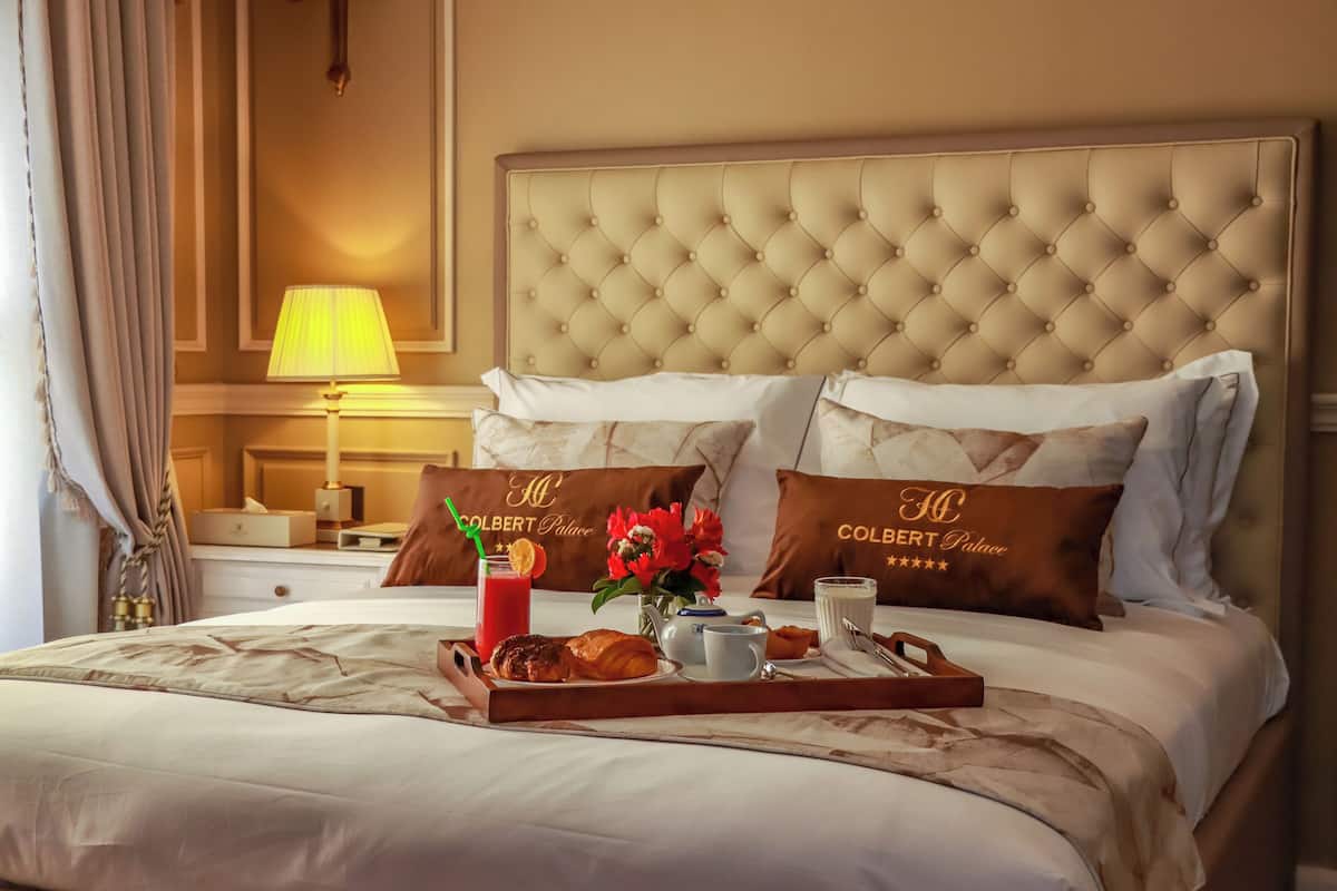 Ein Hotelzimmer mit einer geblümten Kopfteil, zwei Kissen mit dem Hotelnamen, einem Tablett mit Frühstücksangeboten und einem Nachttisch mit einer Lampe.