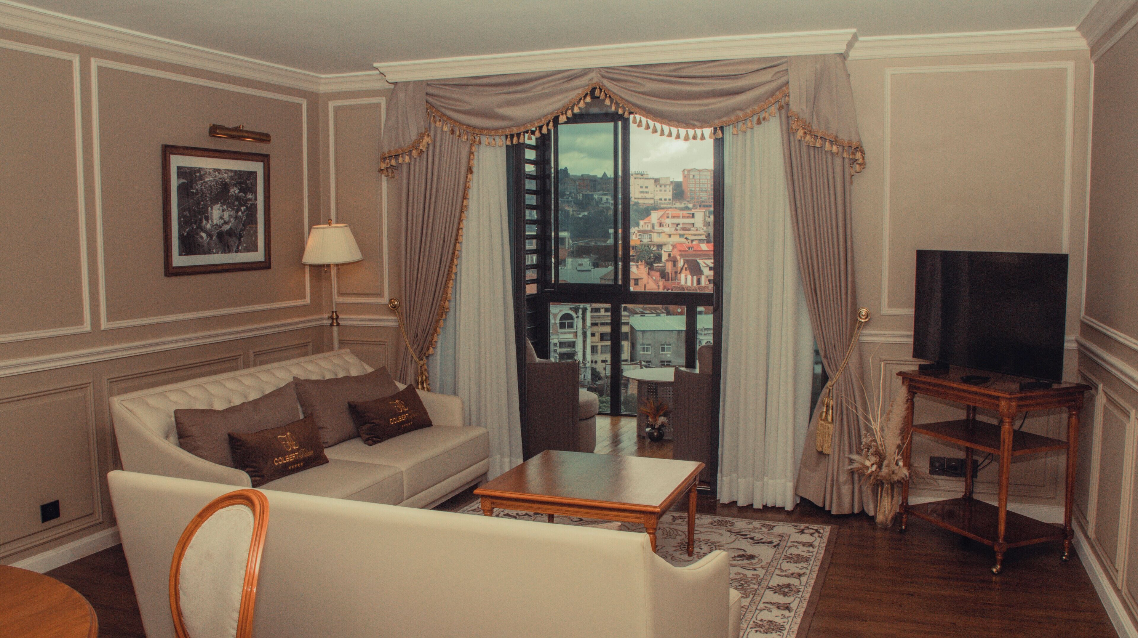 A living room with a sofa, a coffee table, a television on a stand, and a view of buildings through large windows.