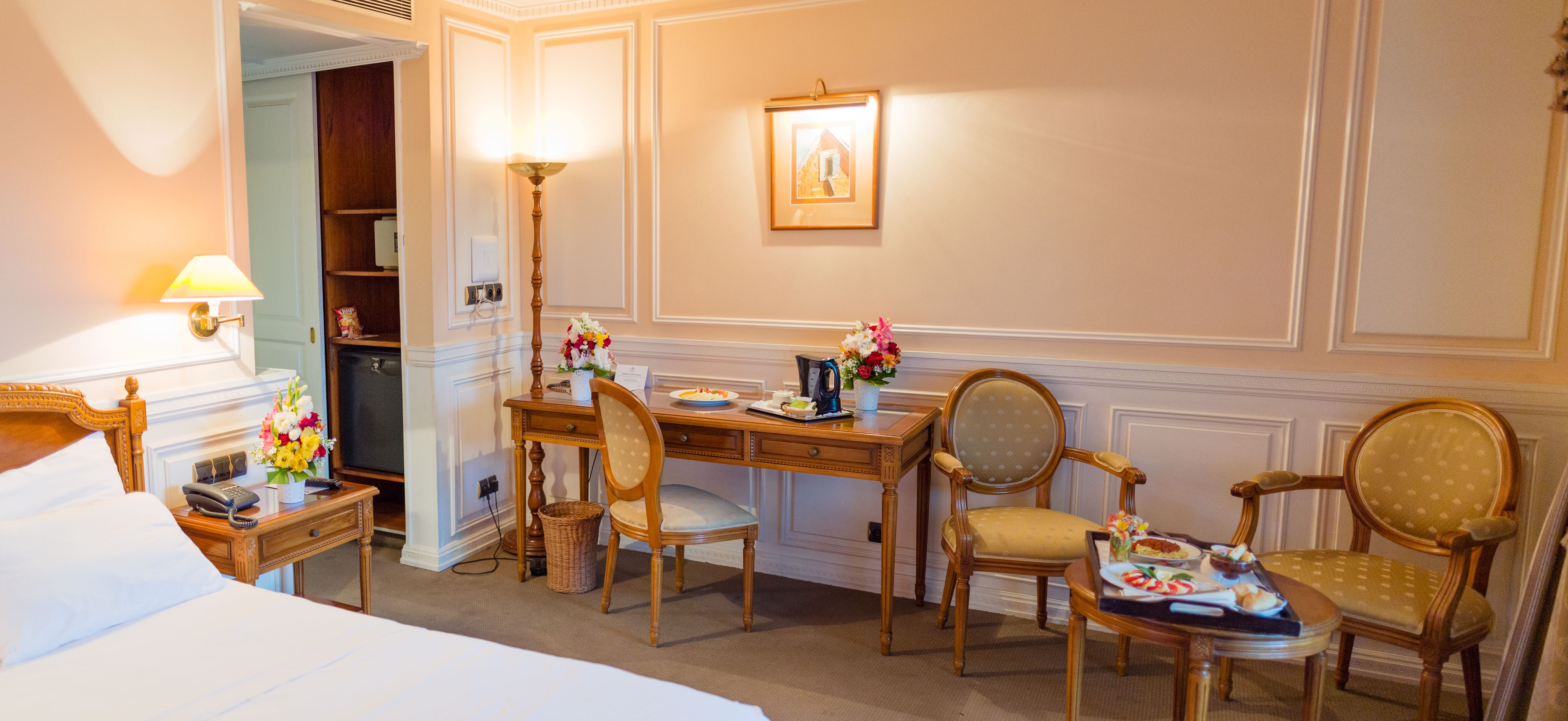 Une chambre d’hôtel avec un lit, un bureau, deux chaises, une lampe, un tableau au mur et une petite table sur laquelle se trouve une assiette de nourriture.