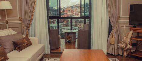 Quarto de hotel com sofá, mesa de centro, televisão e vista para a cidade através de grandes janelas.