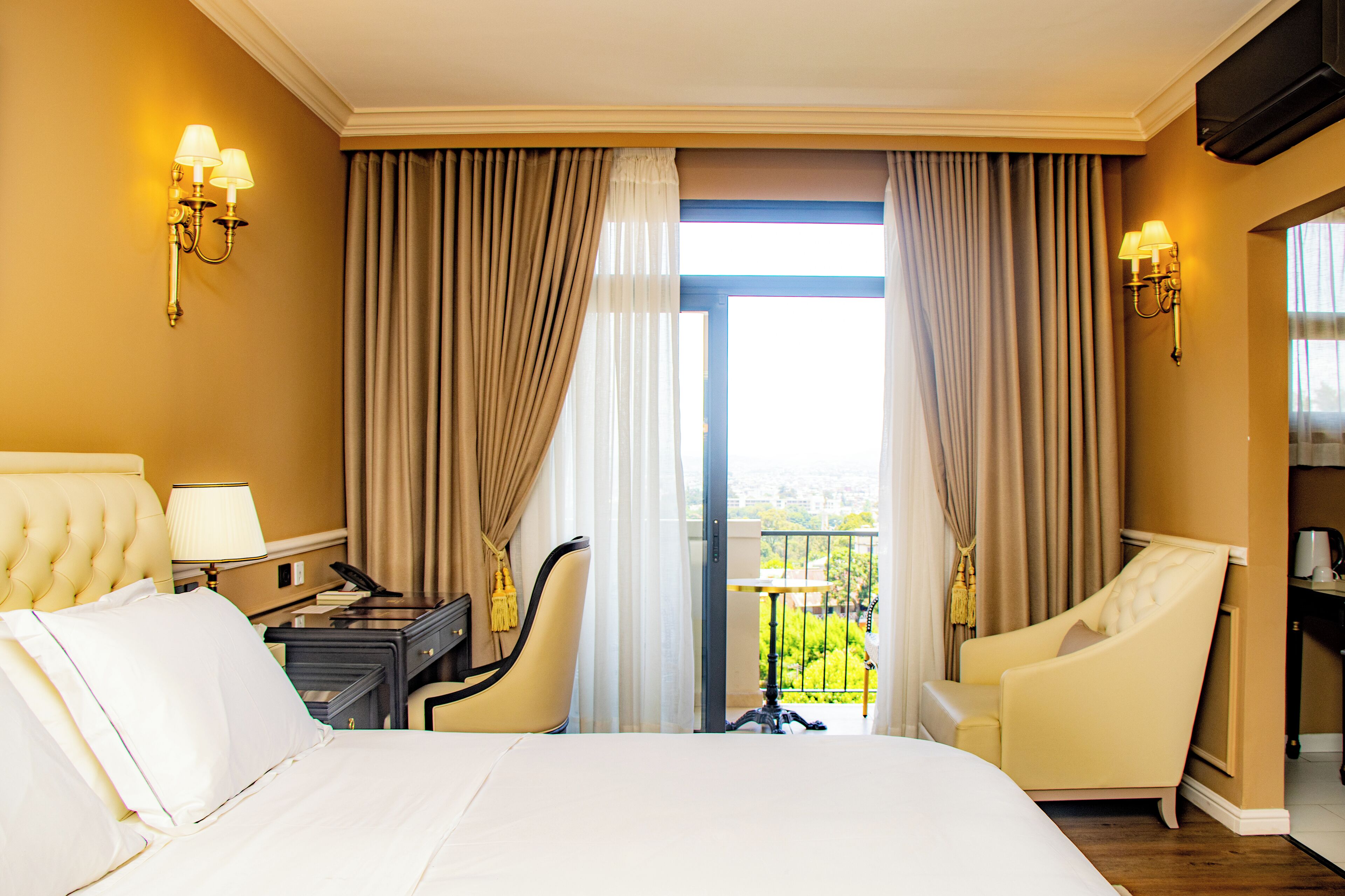 Habitación de hotel con una cama grande, dos sillones, un escritorio con lámpara y un balcón con cortinas.