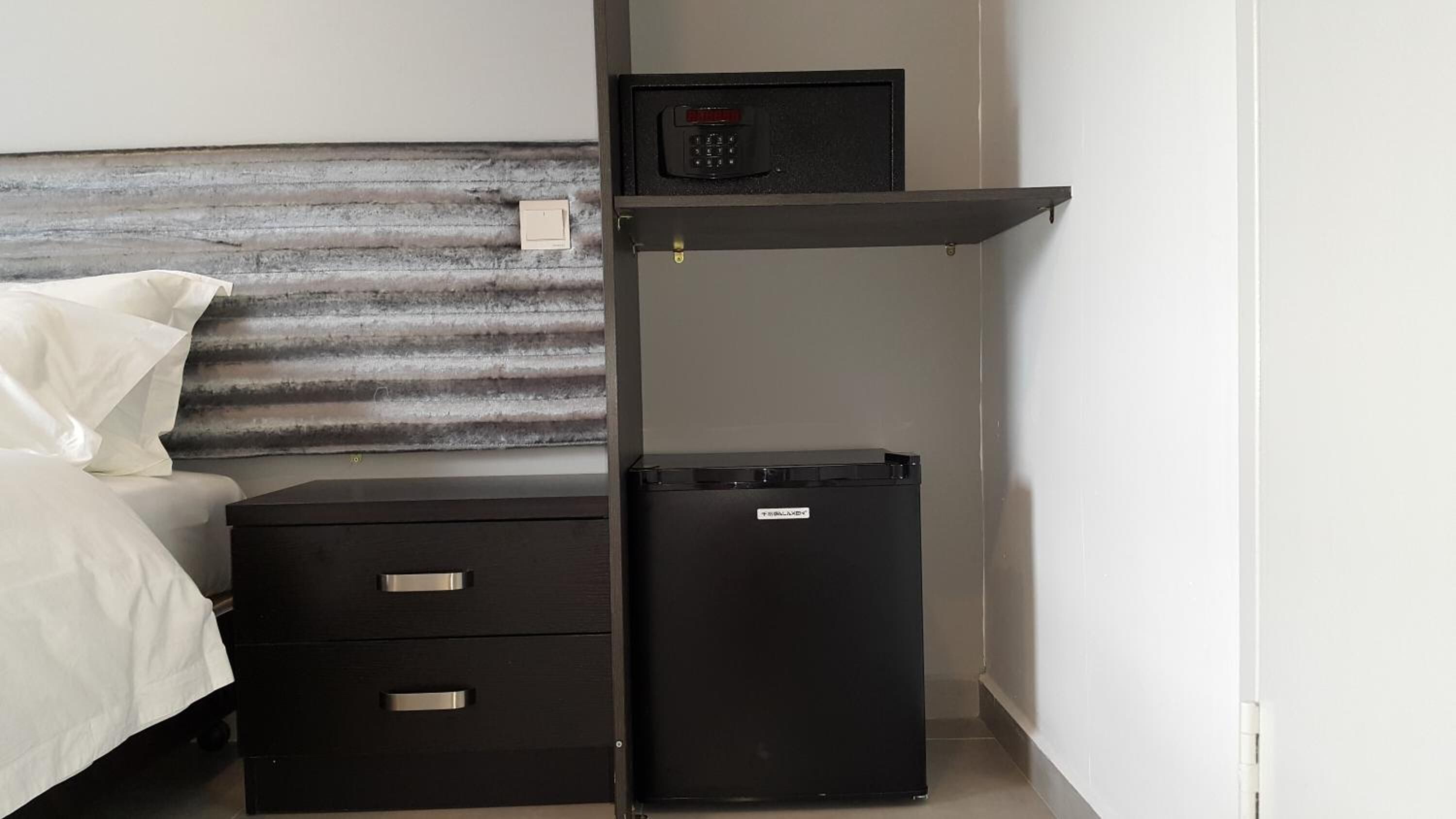 Habitación de hotel compacta con cama, un escritorio pequeño con cajones, un estante con cerradura digital y un refrigerador pequeño.