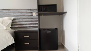 Habitación de hotel compacta con cama, un escritorio pequeño con cajones, un estante con cerradura digital y un refrigerador pequeño.